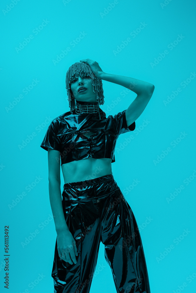 Trendy young woman in latex outfit touching jewelry headwear isolated on blue background with lighting.