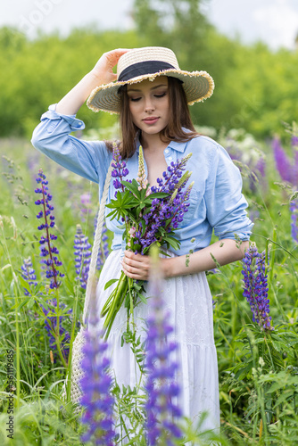 Beautiful young girl in a white dress, straw hat with a bouquet of violet flowers in her hands and picnic basket. Pretty woman in summer in the blooming field holding a bunch of purple lupin
