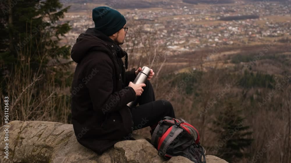 a male tourist with a backpack enjoys the view of the city from the mountain and drinks tea from a thermos a male tourist with a backpack enjoys the view of the city from the mountain and drinks tea from a thermos