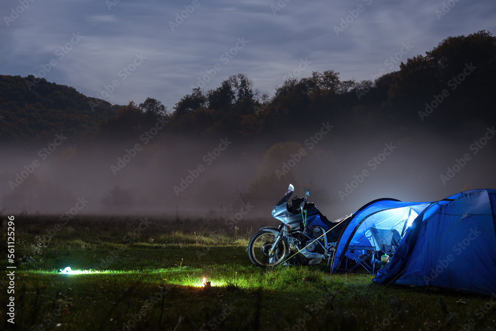 Camping, blue tent with light, touring motorcycle for travel. The concept of recreation and tourism. Space for text, selective focus. Mist, fog and moonlight.