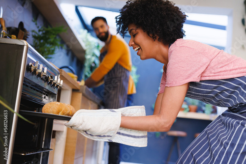 Biracial young man looking at cheerful girlfriend removing freshly baked bread from oven in kitchen