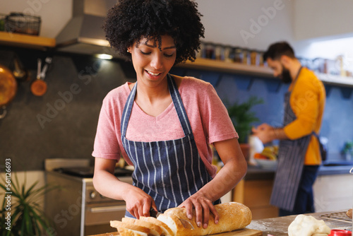 Biracial young woman cutting loaf of bread into slices on table while cooking with boyfriend at home photo
