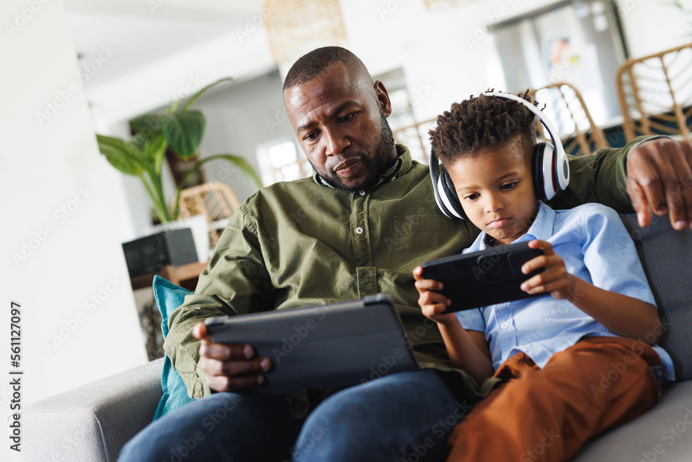 Happy african american father and son sitting on sofa, using tablet and smartphone in living room Happy african american father and son sitting on sofa, using tablet and smartphone in living room