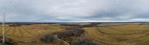 Aerial panorama of spring farmland before crops are planed with a stream surrounded by forest under a sky full of white and gray clouds. 