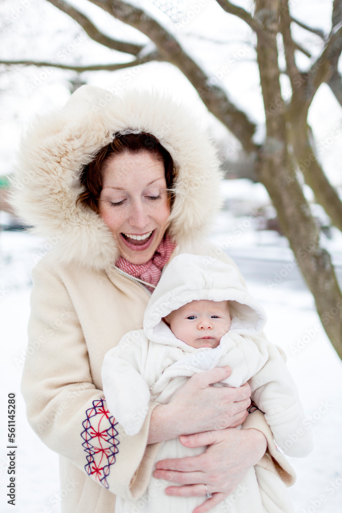 A young mother with red hair in a vintage white coat smiles and holds her newborn baby in a white bunting outside her home in the snow. A young mother with red hair in a vintage white coat smiles and holds her newborn baby in a white bunting outside her home in the snow.