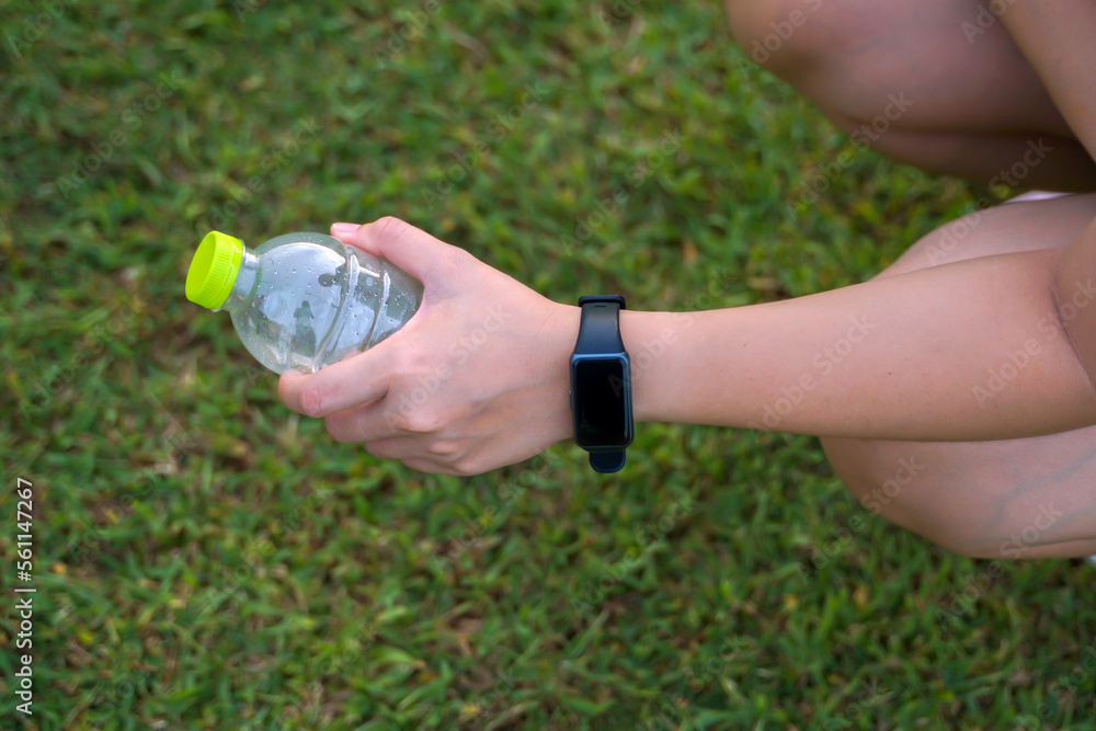 Asian woman wearing smartwatch, holding water bottle, she sits and drinks water during break, in concept, leisure activity, outside, exercise, outdoor activities                          