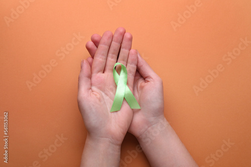 World Mental Health Day. Woman holding green ribbon on pale orange background  top view