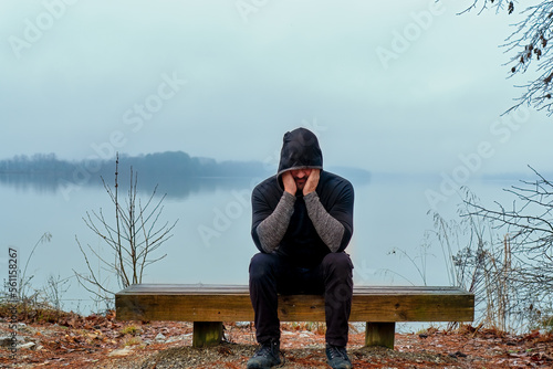 An older depressed man sitting alone on a foggy morning lake bench wearing a black hoodie.