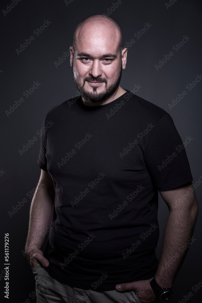 Naklejka premium Smiling bald man with a beard stands on a black background. Brutal guy in a black T-shirt. Vertical.
