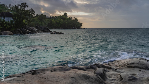 Evening seascape. Granite boulders on the shore of the turquoise ocean. A house is visible among the green vegetation. The sky is highlighted in pink, orange. Seychelles. Mahe.