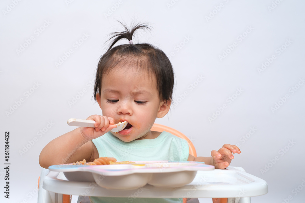 Cheerful baby child sitting and eats food by herself with spoon. Portrait of happy kid boy or girl in high chair. Smiling feeding delicious food concept. Isolate on white background. Cheerful baby child sitting and eats food by herself with spoon. Portrait of happy kid boy or girl in high chair. Smiling feeding delicious food concept. Isolate on white background.