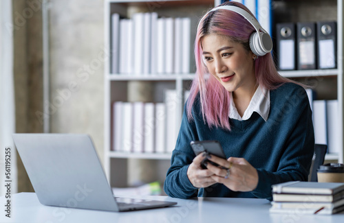 Beautiful Asian businesswoman wearing headphones talking via video call on Her notebook and smartphone to record product information, income, and online business idea.