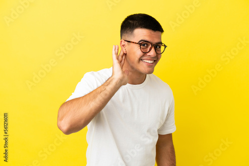 Young handsome man over isolated yellow background listening to something by putting hand on the ear