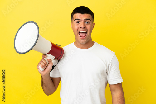 Young handsome man over isolated yellow background holding a megaphone and with surprise expression