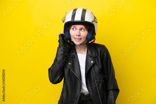 Young English woman with a motorcycle helmet isolated on yellow background listening to something by putting hand on the ear