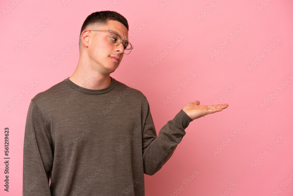Young Brazilian man isolated on pink background holding copyspace with doubts