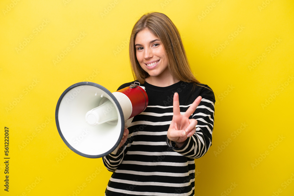 Young caucasian woman isolated on yellow background holding a megaphone and smiling and showing victory sign Young caucasian woman isolated on yellow background holding a megaphone and smiling and showing victory sign