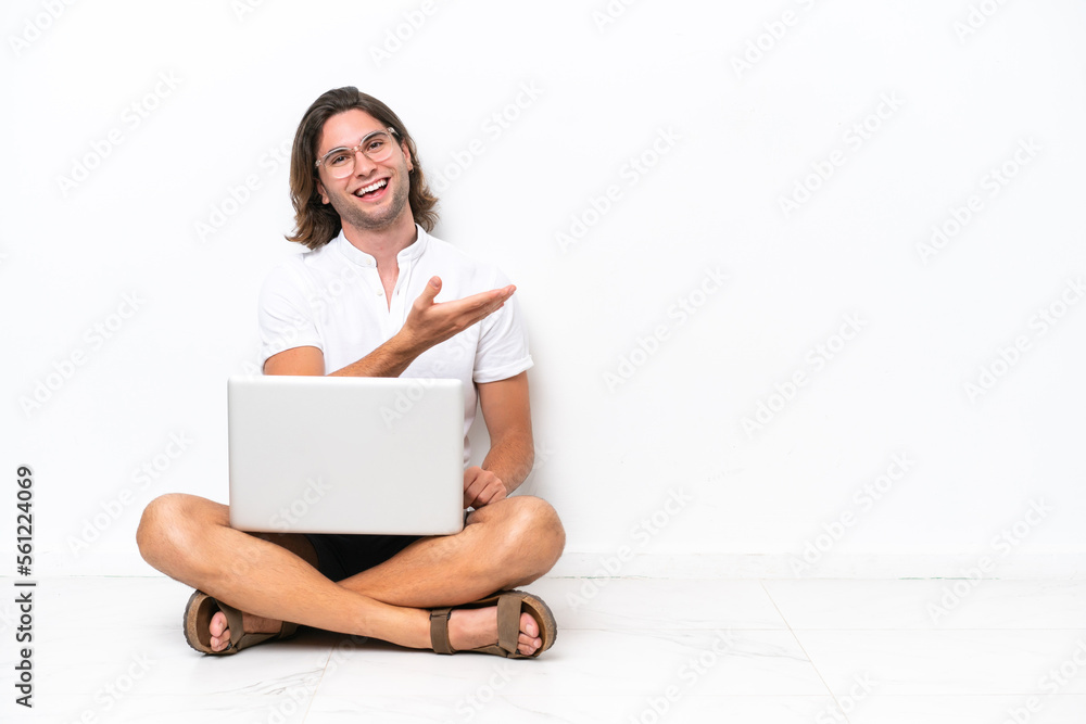 Young handsome man with a laptop sitting on the floor isolated on white background presenting an idea while looking smiling towards Young handsome man with a laptop sitting on the floor isolated on white background presenting an idea while looking smiling towards