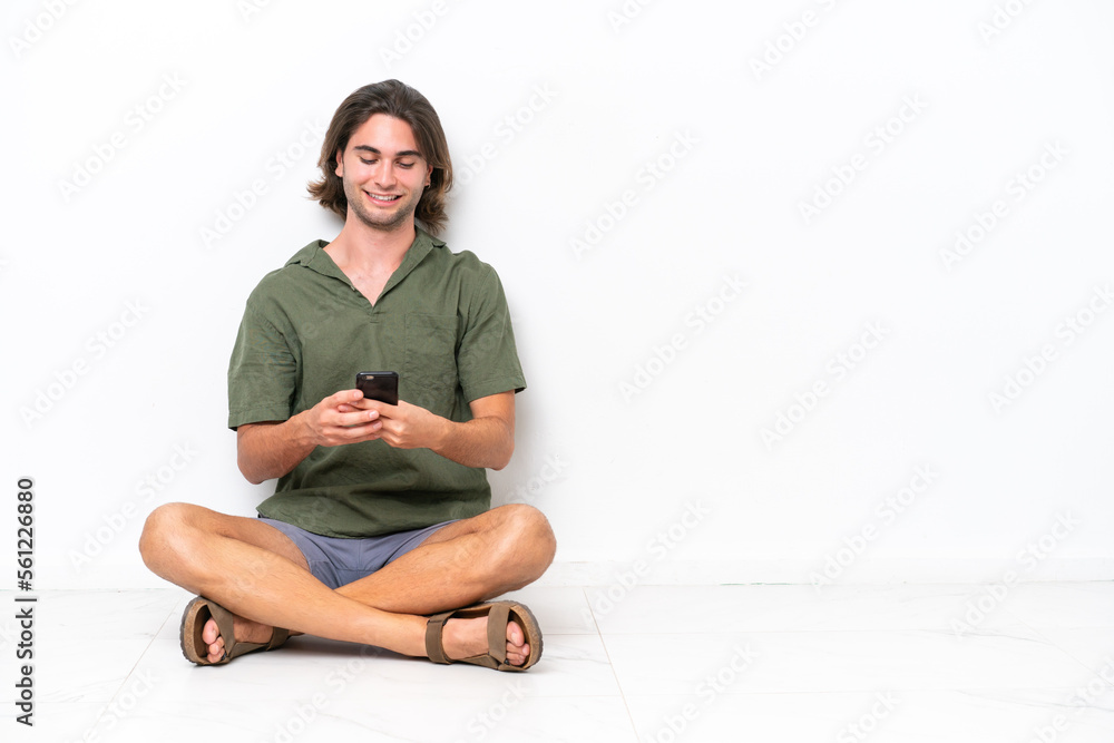 Young handsome man sitting on the floor isolated on white background sending a message with the mobile Young handsome man sitting on the floor isolated on white background sending a message with the mobile