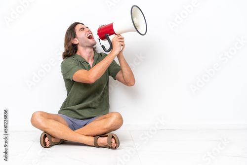 Young handsome man sitting on the floor isolated on white background shouting through a megaphone