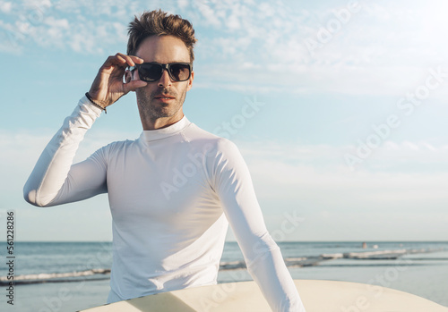 Konstfotografi Young handsome surfer guy wearing an empty blank lycra t-shirt and holding a surfboard