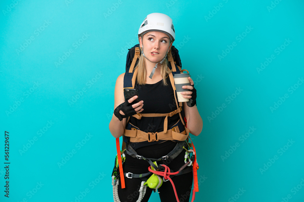 Young caucasian rock climber woman isolated on blue background holding coffee to take away and a mobile while thinking something Young caucasian rock climber woman isolated on blue background holding coffee to take away and a mobile while thinking something