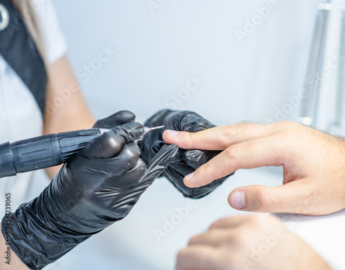 Manicurist master is doing hardware manicure to man  hands close-up. She is removing cuticle and pterygium on nail using electric apparatus. Hygiene and care for hands. Beauty industry concept