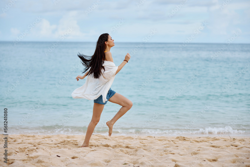 Sports woman runs along the beach in summer clothes on the sand in a yellow tank top and denim shorts white shirt flying hair ocean view, beach vacation and travel Sports woman runs along the beach in summer clothes on the sand in a yellow tank top and denim shorts white shirt flying hair ocean view, beach vacation and travel