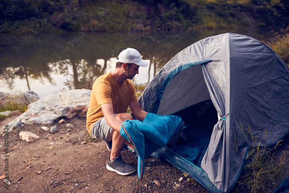 Man camping in nature, setting up the tent for overnight staying near forest river.