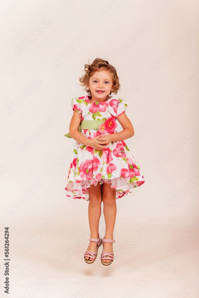 Full length fashionable little girl model jumping in stylish flowers sundress on white background, looking at camera. Studio shot child in sundress. Fashion trendy concept. Copy text space for ad
