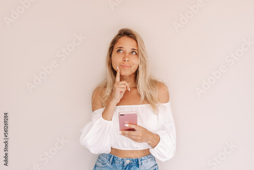 A young caucasian attractive thoughtful blonde woman holds mobile phone in her hand and thinks what to order looking up at empty copy space for text or design isolated on a beige background
