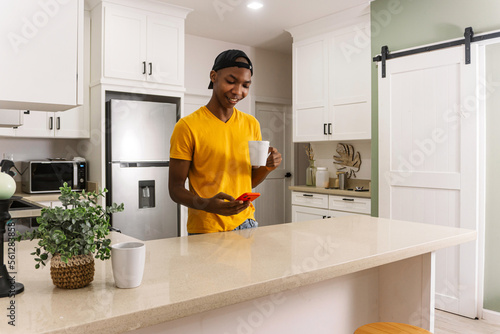 Young african man in the kitchen with a cup of coffee and using a phone.