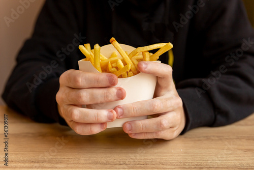 large hands of a man in a black hoodie with vitiligo to hold French fries in paper packaging in a cafe at the table during the day, fast food at lunch with coffee fingers wrapped around a paper eco