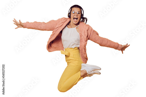 Fotografiet Excited brunette girl in glasses jumping, listening music in headphones spread out her arms dressed in yellow pants, white shoes, pink sweater and headphones on her head over transparent background
