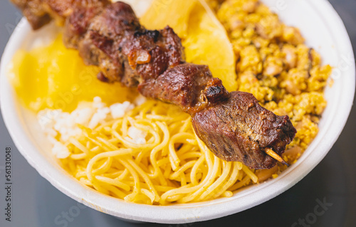 Churrasquinho brasileiro. Espetinho com carne bovina sobre uma marmita de isopor com arroz, feijão tropeiro, mandioca cozida e macarrão. Comida brasileira, churrasco.