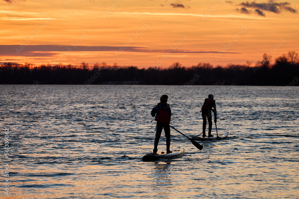 Black sunset silhouettes of two young athletes paddling on stand up paddleboard at river at sunset in cold season. Healthy lifestyle.