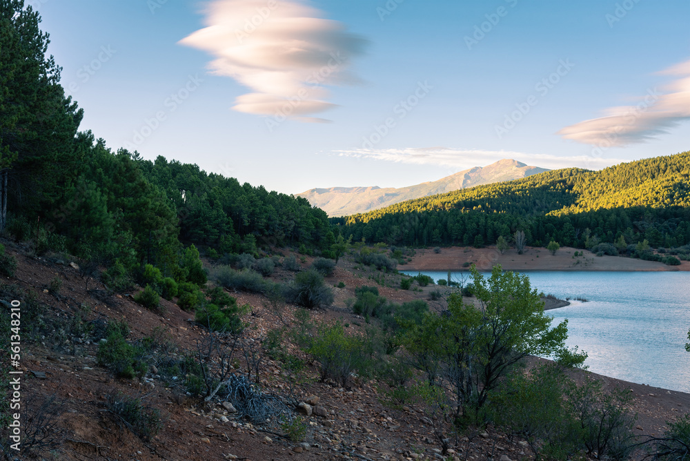 Fototapeta premium Beautiful landscape of a lake surrounded by greenery mountains and trees under a blue sky with amazing clouds at sunset, El Vado, Guadalajara, Spain