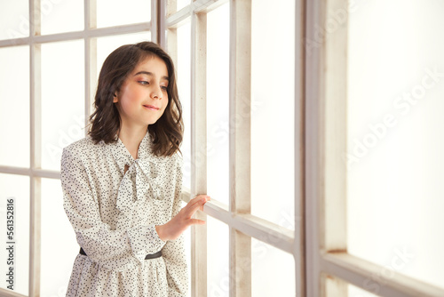 Portrait little girl in stylish white dress posing near large light window in living room, looking away. Studio shot smiling positive teen lady. Child emotion concept. Copy text space for advertising