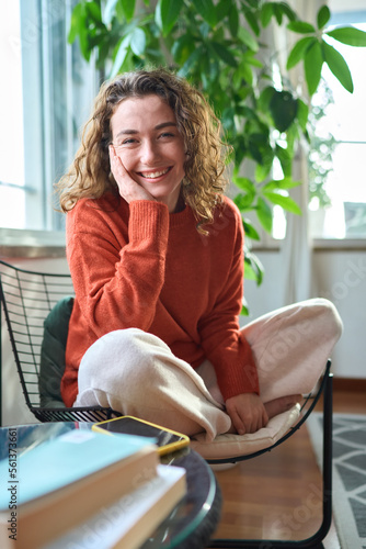 Happy pretty young positive woman sitting on chair relaxing at home looking at camera. Smiling cheerful lady chilling in apartment, laughing, feeling relaxed and satisfied. Vertical portrait.