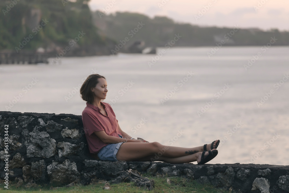 Thoughtful woman sitting on retaining wall by sea during sunset Thoughtful woman sitting on retaining wall by sea during sunset