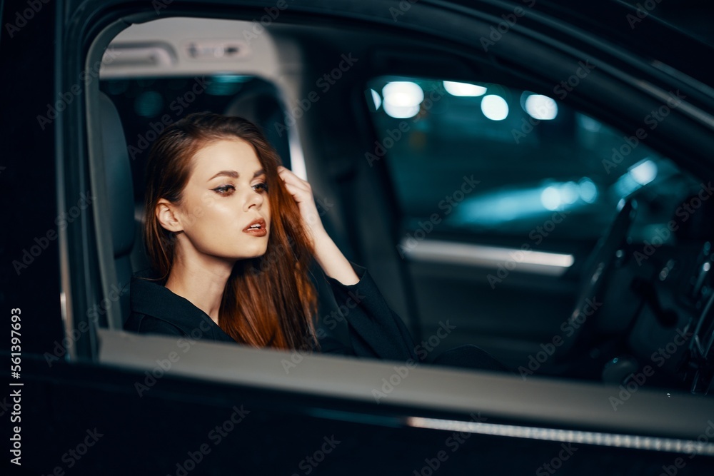 a stylish, luxurious woman sits in a black car at night, touching her face with a thoughtful expression. Close horizontal photo