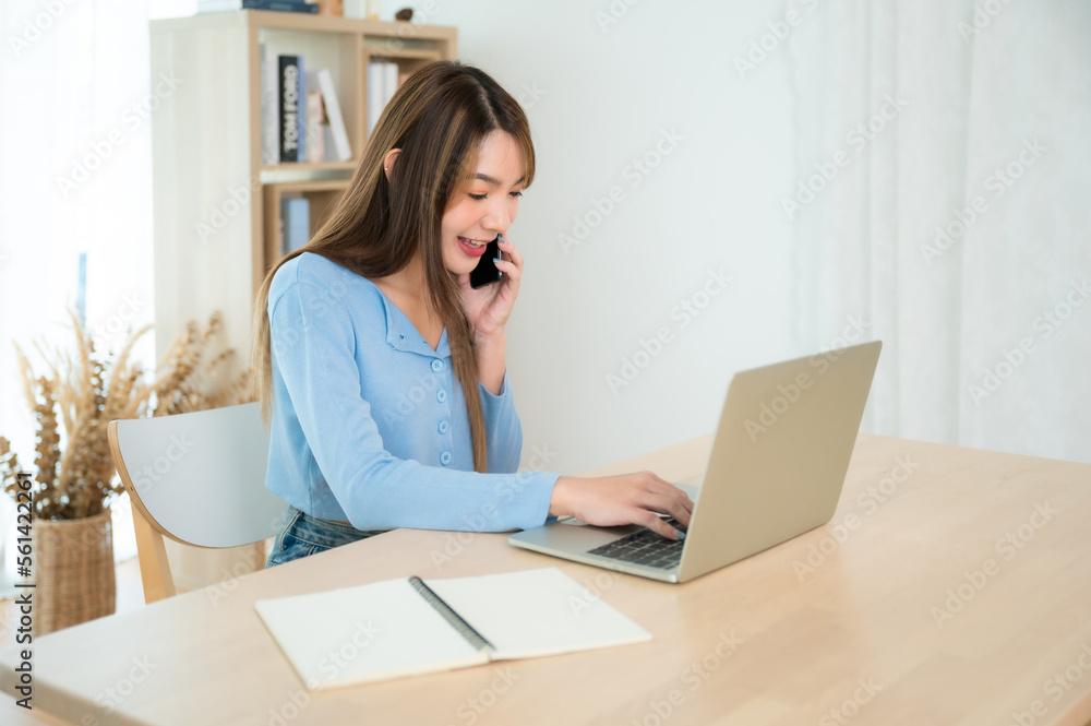 Young asian woman talking mobile smartphone while working on computer laptop on desk in living room at home Young asian woman talking mobile smartphone while working on computer laptop on desk in living room at home