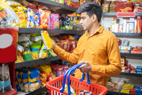 Young Indian man buying grocery at shop or supermarket. Asian male choose snacks and food items while holding basket.