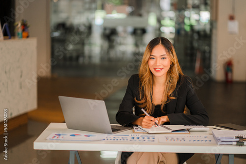 Asian businesswoman working on charts and graphs showing results in office with documents and laptop Worker documents calculating financial indicators, smiling and happy with success.