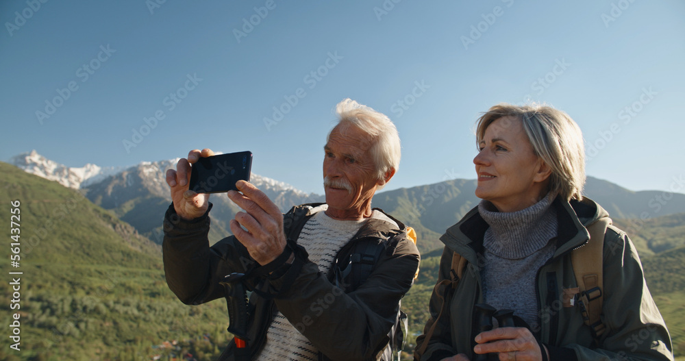 Senior caucasian couple having a nordic walk in mountains, then stopping to take a picture with smartphone. Old people travelling together after retirement - pension, tourism concept Senior caucasian couple having a nordic walk in mountains, then stopping to take a picture with smartphone. Old people travelling together after retirement - pension, tourism concept