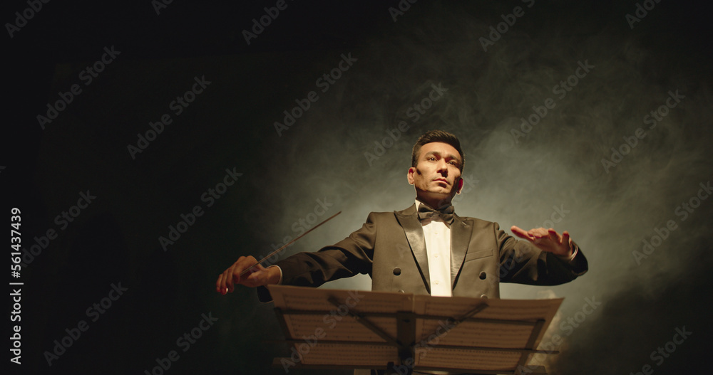 Asian symphony orchestra conductor wearing suit is directing musicians with movement of baton, isolated on black smokey background Asian symphony orchestra conductor wearing suit is directing musicians with movement of baton, isolated on black smokey background