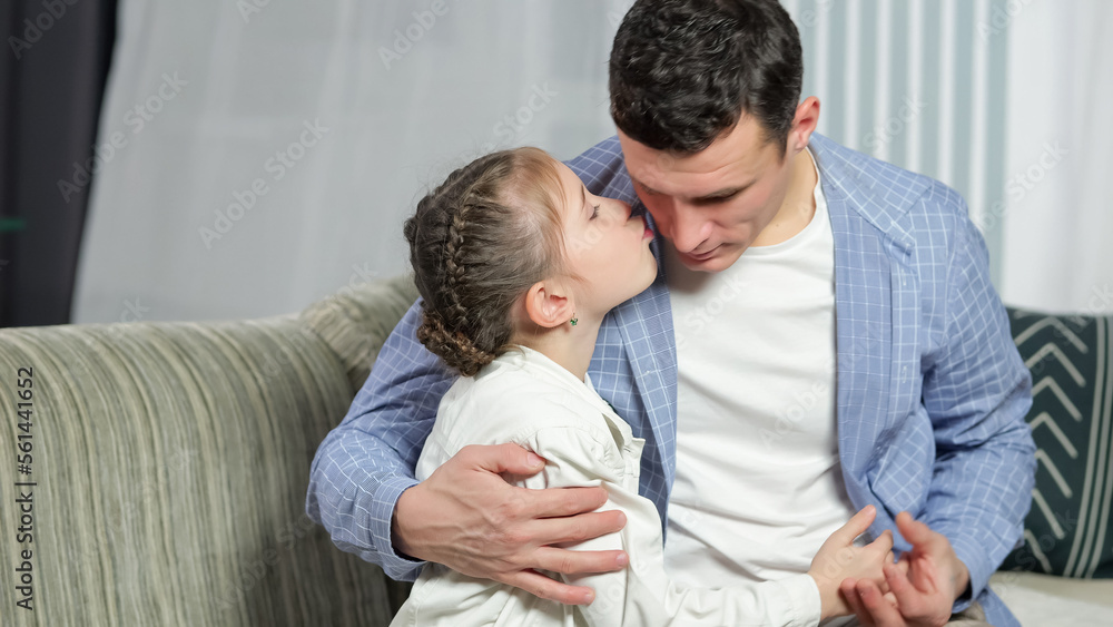 Loving father shows love and embraces precious daughter at home. Lovely junior schoolgirl kisses father on cheek and hugs man tight on sofa in living room Loving father shows love and embraces precious daughter at home. Lovely junior schoolgirl kisses father on cheek and hugs man tight on sofa in living room