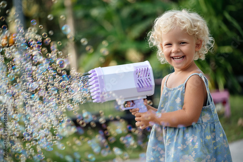 Portrait of a Child of a curly blonde girl happily playing with soap bubbles from a large air gun. Outdoors in summer.