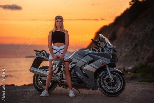 Alone caucasian woman posing at the beach holds a helmet. Golden sunset and motorbike on the background. Concept of World Motorcyclist Day and moto travel