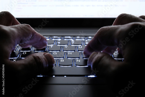 Man working on a laptop with hands typing on a backlit keyboard in the dark late at night. Hacker secretly accessing company files, stealing government data. Concept for corporate espionage, hacking.